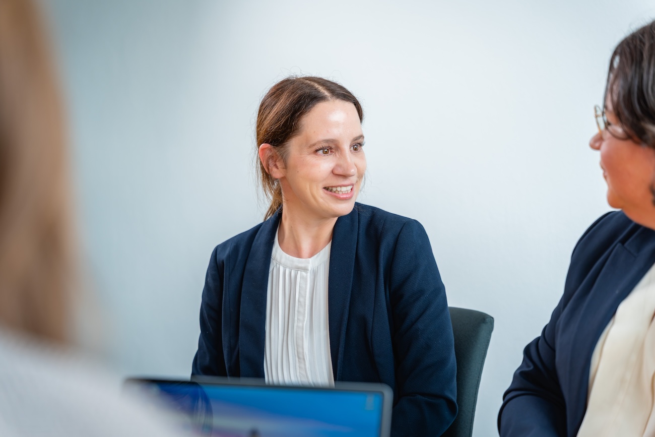 Zwei Frauen in Businesskleidung führen ein Gespräch in einem hellen Raum, wobei die Frau links lächelnd zur anderen Frau schaut.