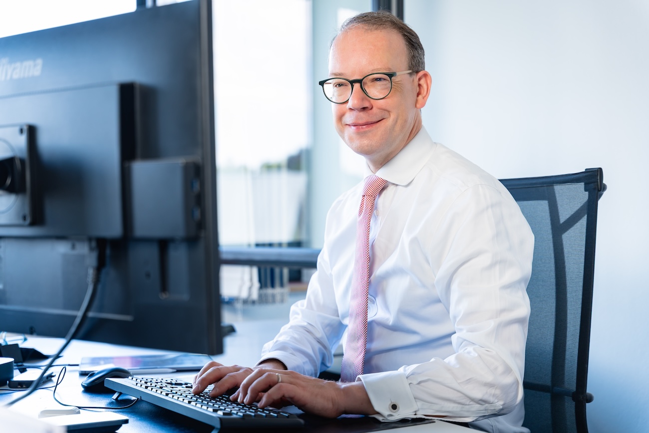 Ein lächelnder Mann mit Brille sitzt an einem Schreibtisch und tippt auf einer Tastatur vor einem großen Monitor in einem hellen Büro.