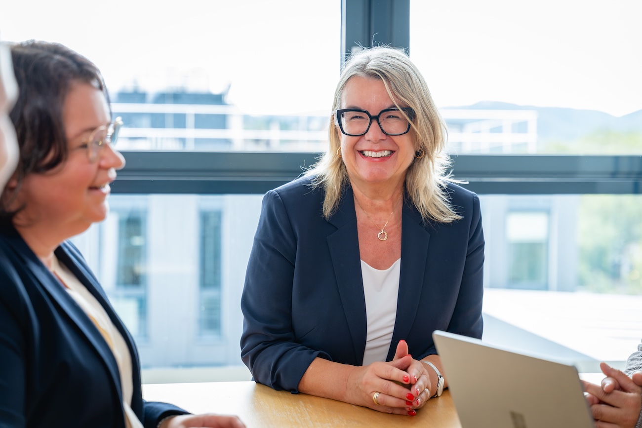 Lächelnde blonde Frau mit Brille in einem dunklen Blazer sitzt an einem Holztisch in einem hellen Büro mit Fenster im Hintergrund, neben ihr eine weitere Frau und ein Laptop.