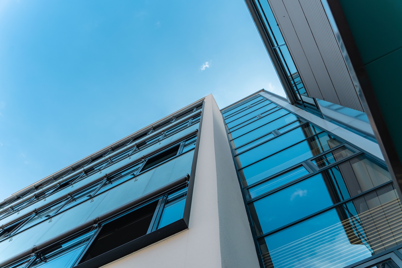 Blick nach oben auf eine moderne Gebäudeecke mit blau getönten Glasfenstern und klarem Himmel im Hintergrund.