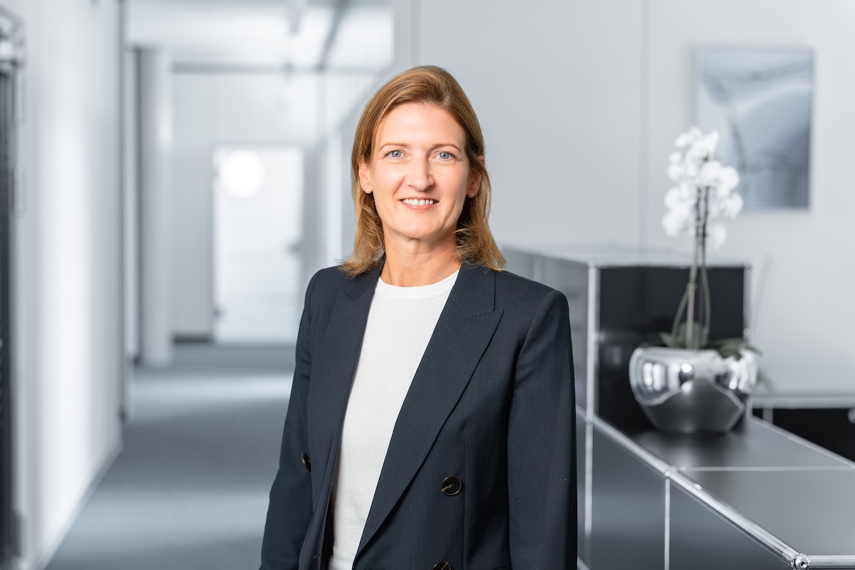 Frau mit mittellangen hellbraunen Haaren trägt einen dunklen Blazer über einem weißen Shirt und lächelt in einem hellen, modernen Büroflur mit schwarzen Möbeln und einer weißen Orchidee in einer silbernen Vase.