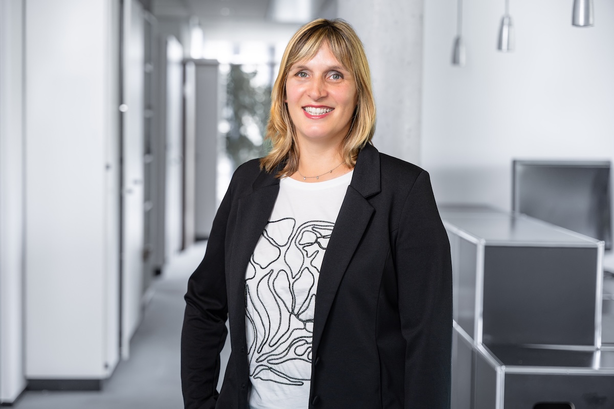 Frau mit blonden Haaren, die in einem schwarzen Blazer und einem weißen T-Shirt mit schwarzem abstraktem Muster in einem modernen Büro lächelt.