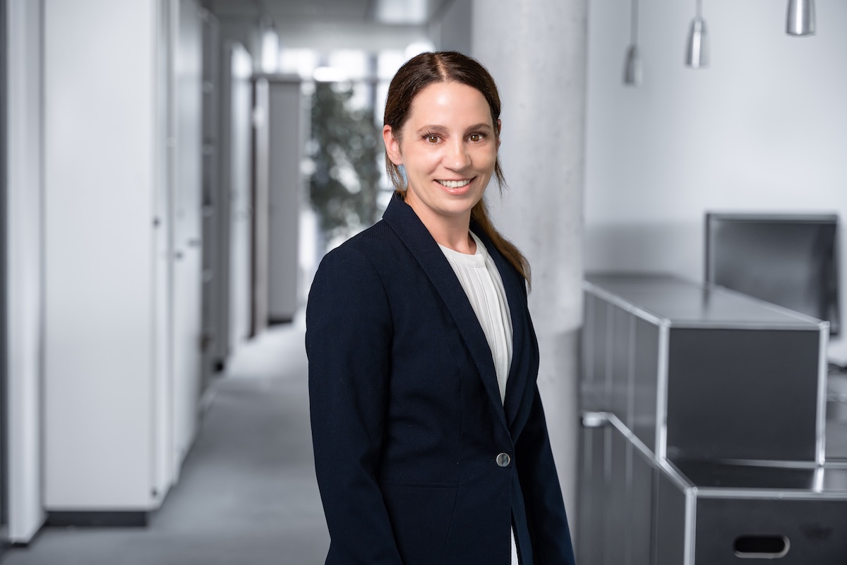 Eine Frau mit dunkelbraunem, zurückgebundenem Haar trägt einen schwarzen Blazer und ein weißes Oberteil, sie steht in einem modernen Büro mit hellen Wänden und Möbeln und lächelt in die Kamera.