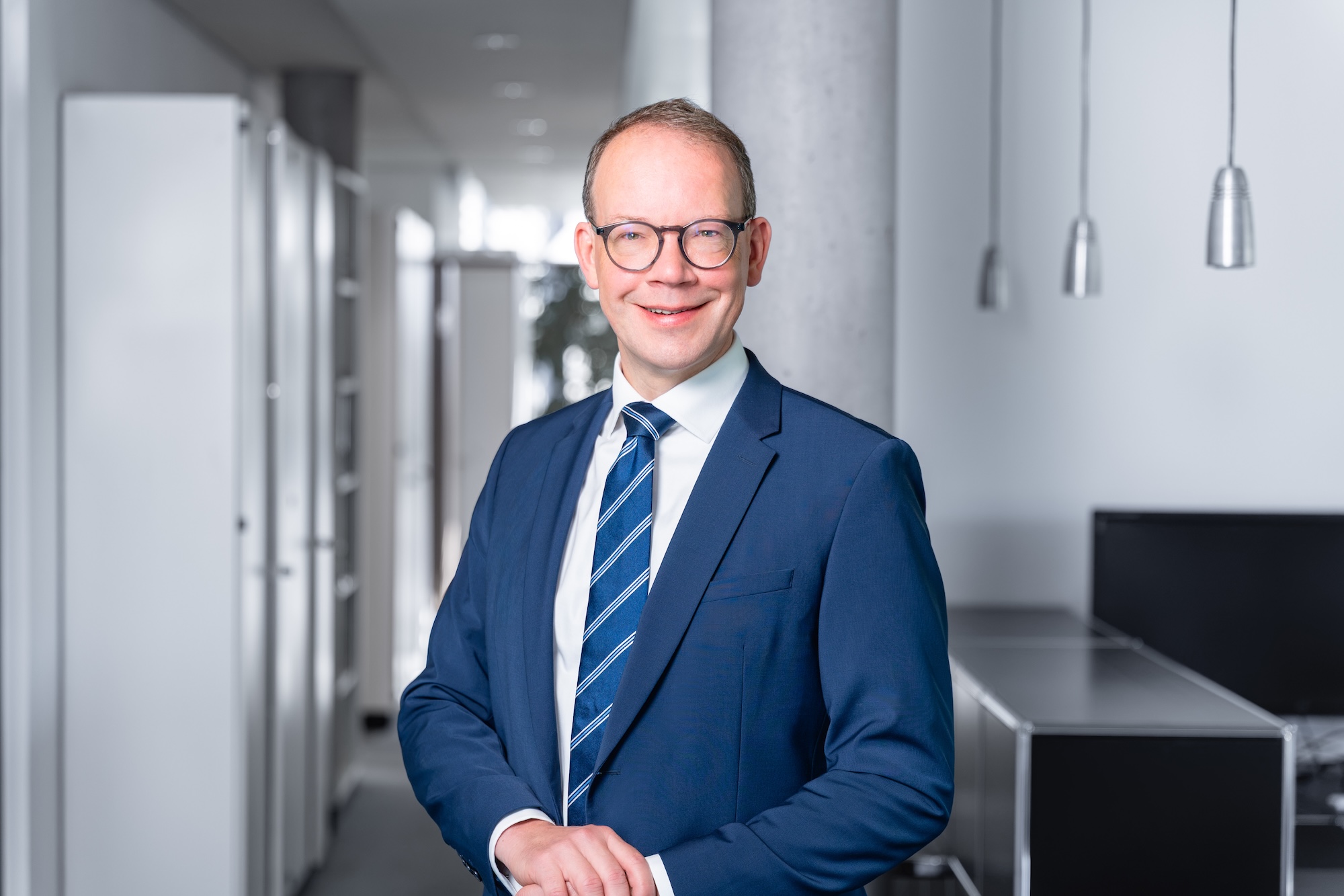 Porträt eines lächelnden Mannes in einem blauen Anzug, weißem Hemd und gestreifter Krawatte vor einem modernen Büroflur mit Betonpfeiler und Hängelampen.