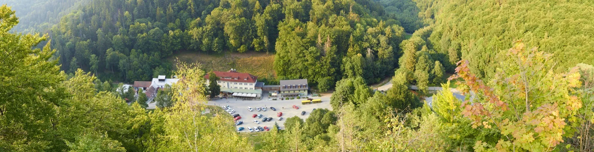 Záběr z výšky na parkoviště s několika auty a budovy obklopené hustým zeleným lesem v horské oblasti.