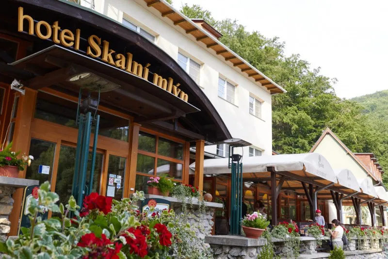 Vstup do hotelu Skalní mlýn s květinovou výzdobou a zeleným zalesněným pozadím.