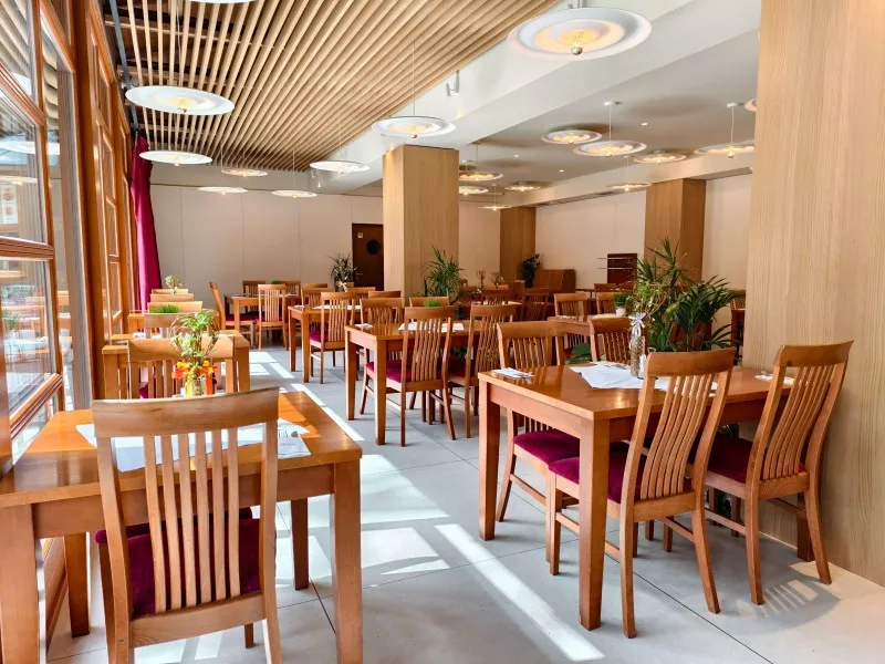 Světla slunce prosvítají okny do moderní restaurace s dřevěnými stolky a židlemi s červenými sedáky a stropem z dřevěných lamel.