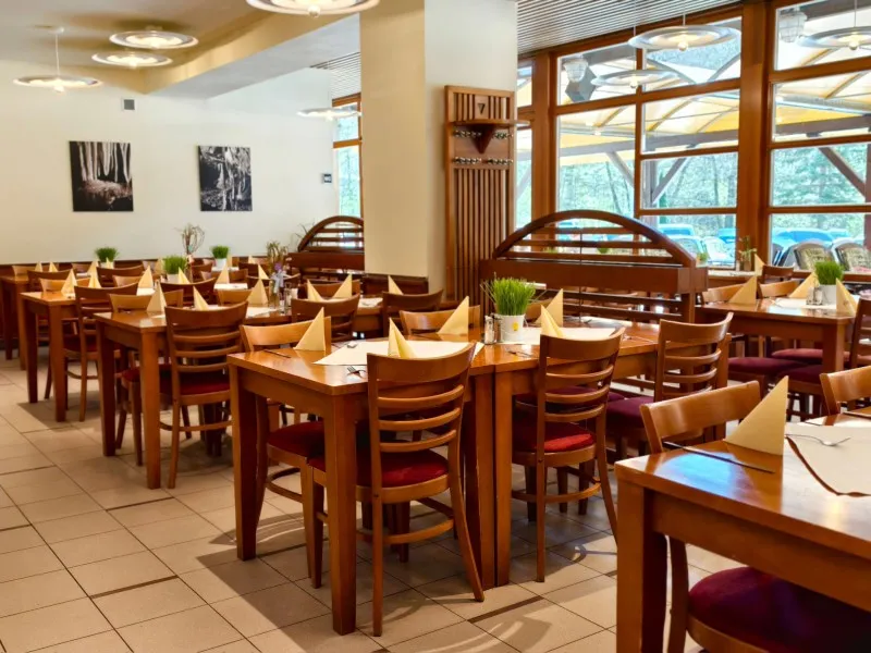 Restaurace s dřevěnými stoly a židlemi, prostřenými ubrousky ve tvaru trojúhelníků a zelenými dekoracemi.
