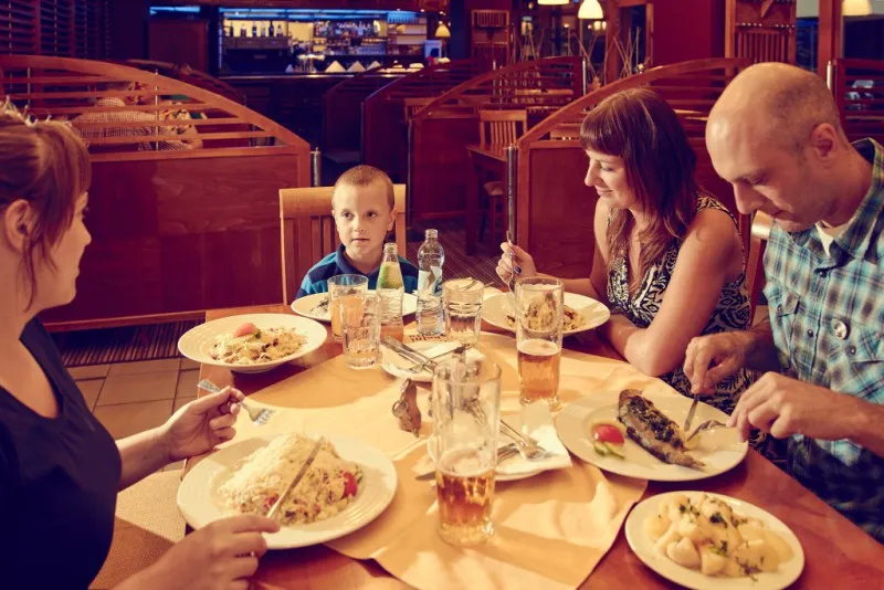 Rodina se čtyřmi lidmi sedí u stolu v restauraci a jí jídlo zařízené dřevěným nábytkem.