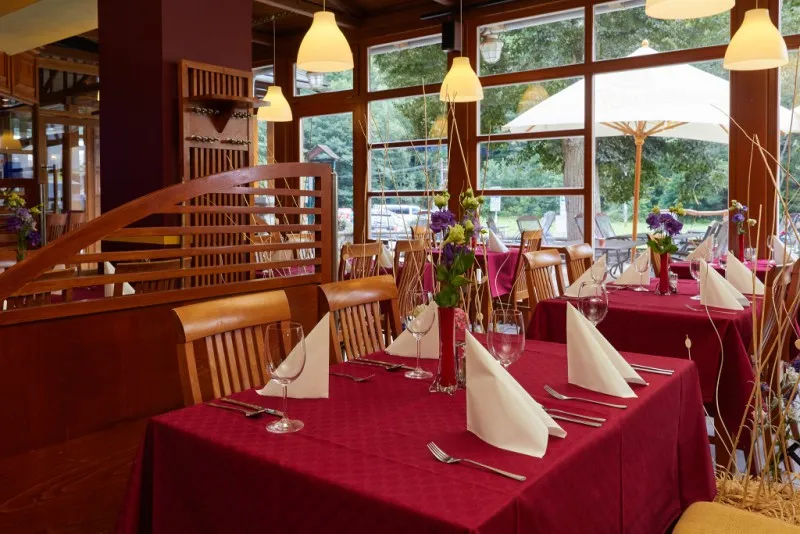 Restaurace s červenými ubrusy na stolech, sklopenými bílými ubrousky a vázičkami s květinami u velkých oken s výhledem na venkovní posezení s bílým slunečníkem.