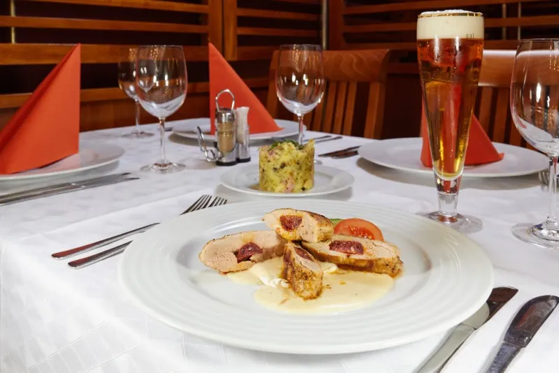 Talíř s kuřecím masem plněným sušenými rajčaty, bramborový salát a sklenice piva na prostřeném stole v restauraci.