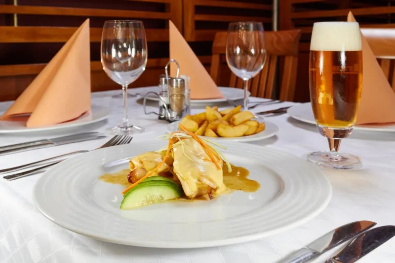 Talíř s pokrmem z kuřecího masa s omáčkou, plátky okurky a hranolky na stole v restauraci vedle sklenice piva s pěnou.