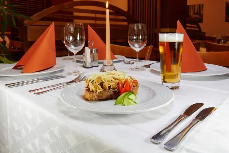 Elegant restaurant table set with white tablecloth, two knives and forks, two wine glasses, a lit candle, orange folded napkins, a plate with open sandwich topped with cheese and vegetables, and a glass of beer.