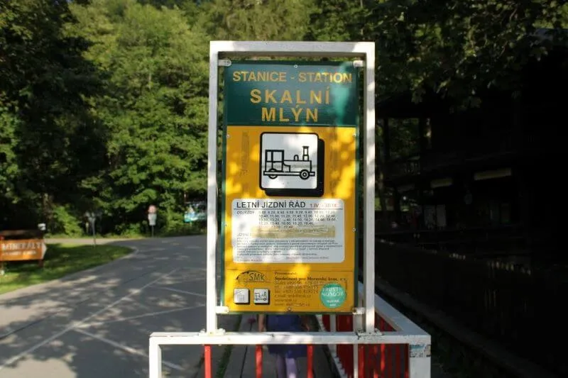 Známka stanice Skalní Mlýn s jízdním řádem a symbolem vlaku na žlutém pozadí u cesty vedoucí do lesa.
