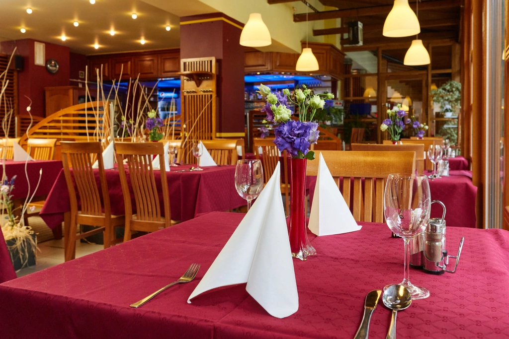 Restaurace s dřevěným nábytkem, ubrusy vínové barvy, sklopenými bílými ubrousky a vázičkami s fialovými květinami na stolech.
