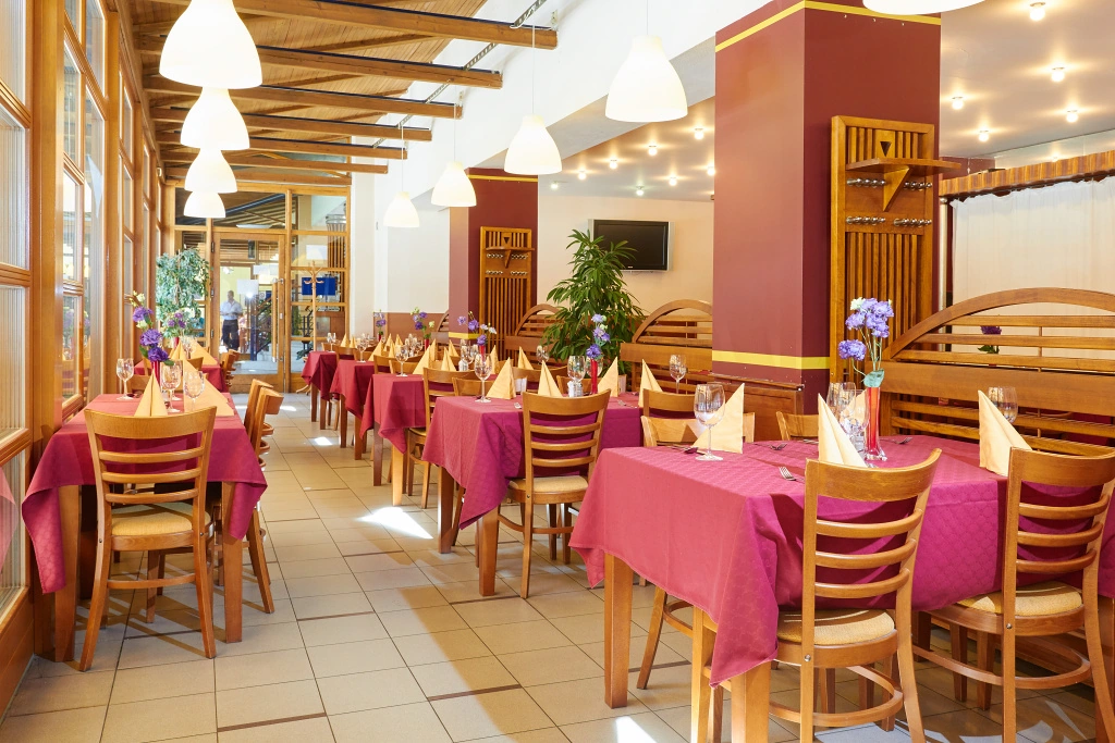 Restaurace s dřevěným nábytkem, vínově červenými ubrusy na stolech a žlutě složenými ubrousky.