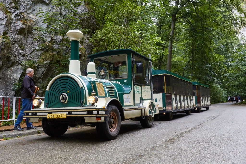 Turistický vláček Eko Expres stojící na silnici obklopené zelenými stromy a skalní stěnou.