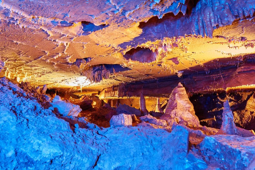 Podzemní jeskyně s barevným osvětlením, stalaktity visící ze stropu a stalagmity vyrůstající ze země.