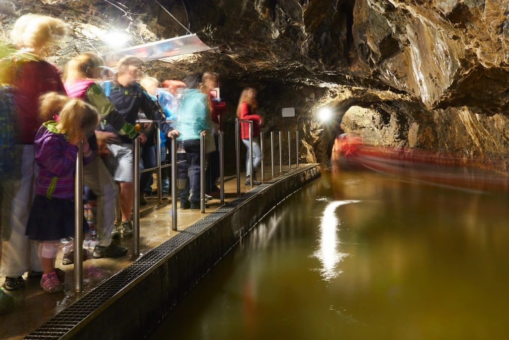 Skupina lidí stojící na plošině v povrchové jeskyni nad podzemní řekou s lodí plující v tunelu.