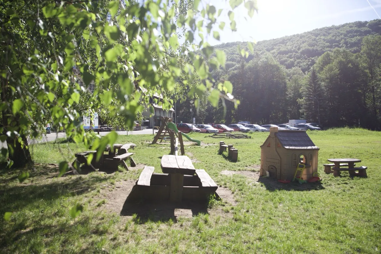 Dětské hřiště s dřevěnými lavicemi, domečkem, skluzavkou a auty na pozadí v přírodě.