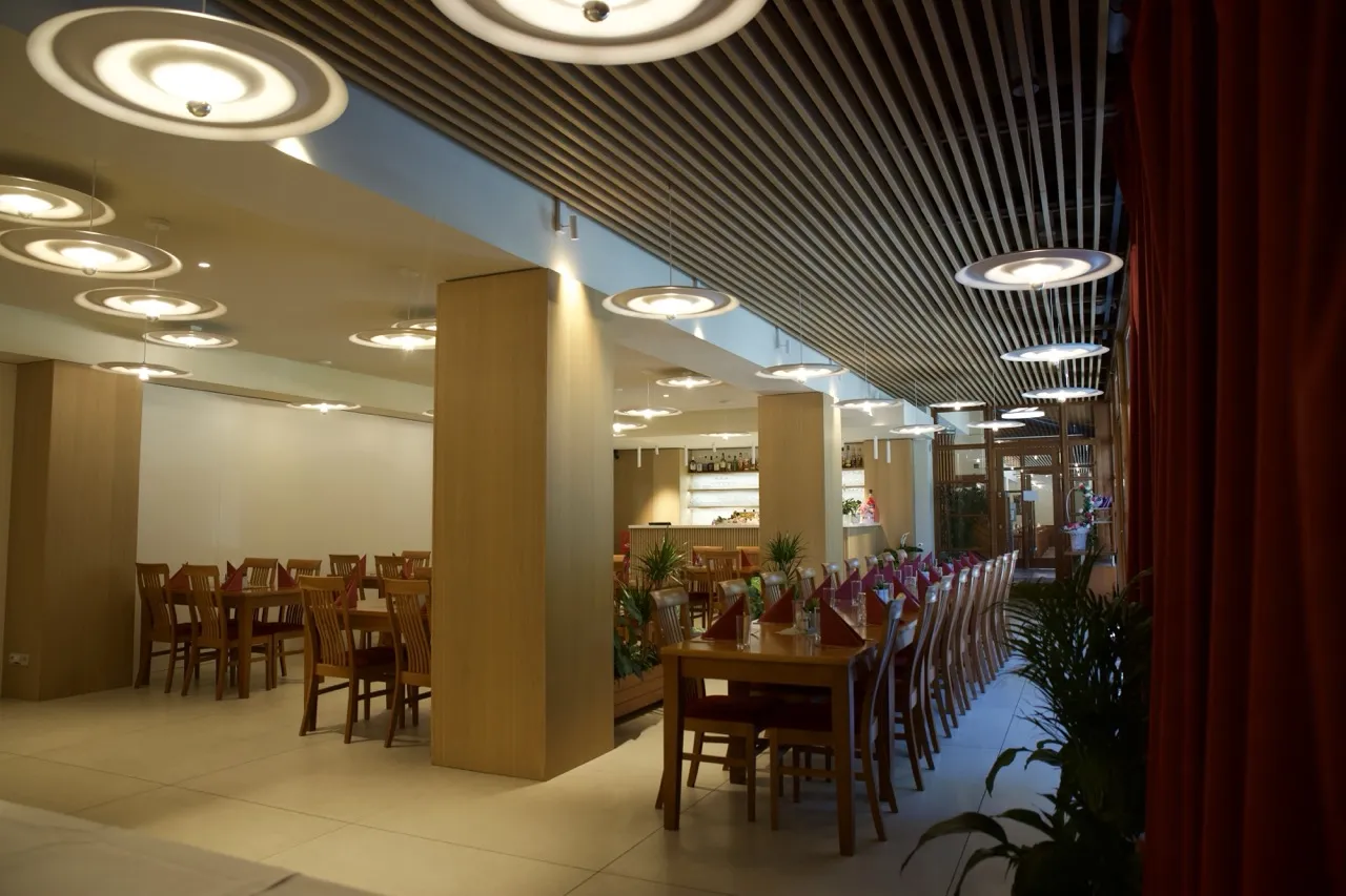 Restaurace s dlouhým stolem, židlemi, závěsnými kruhovými světly a dřevěnými sloupy.