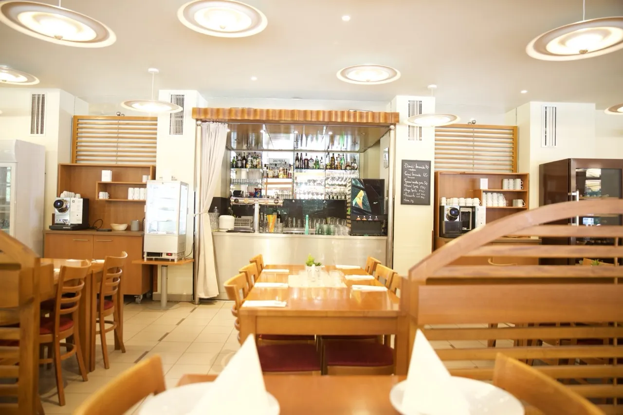 Interiér světlé restaurace s dřevěným nábytkem a barem se zrcadlem a nápoji.