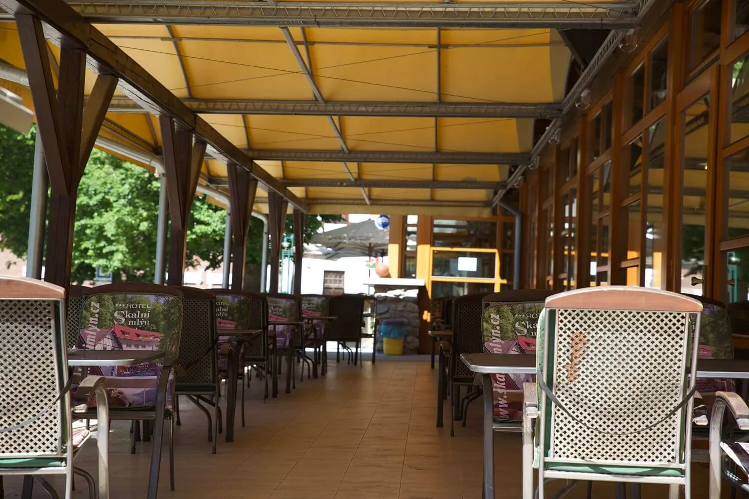 Letní terasa s prázdnými židlemi a stolky pod žlutým zastřešením u hotelu.