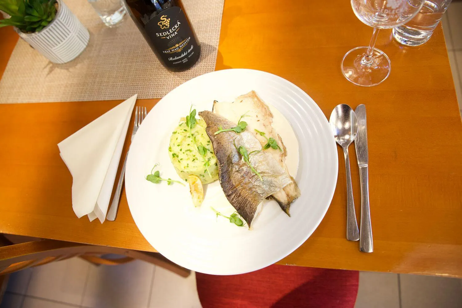 Talíř s pečenou rybou a bramborovou kaší, láhev vína a sklenice na stole v restauraci.
