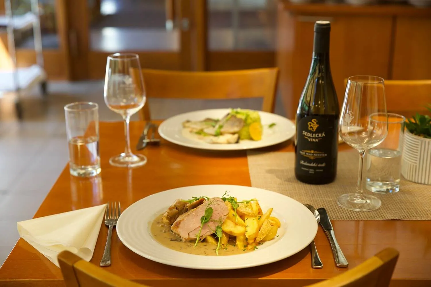 Jídlo v hotelu Skální mlýn s talíři, lahví vína a sklenicemi na stole v restauraci.
