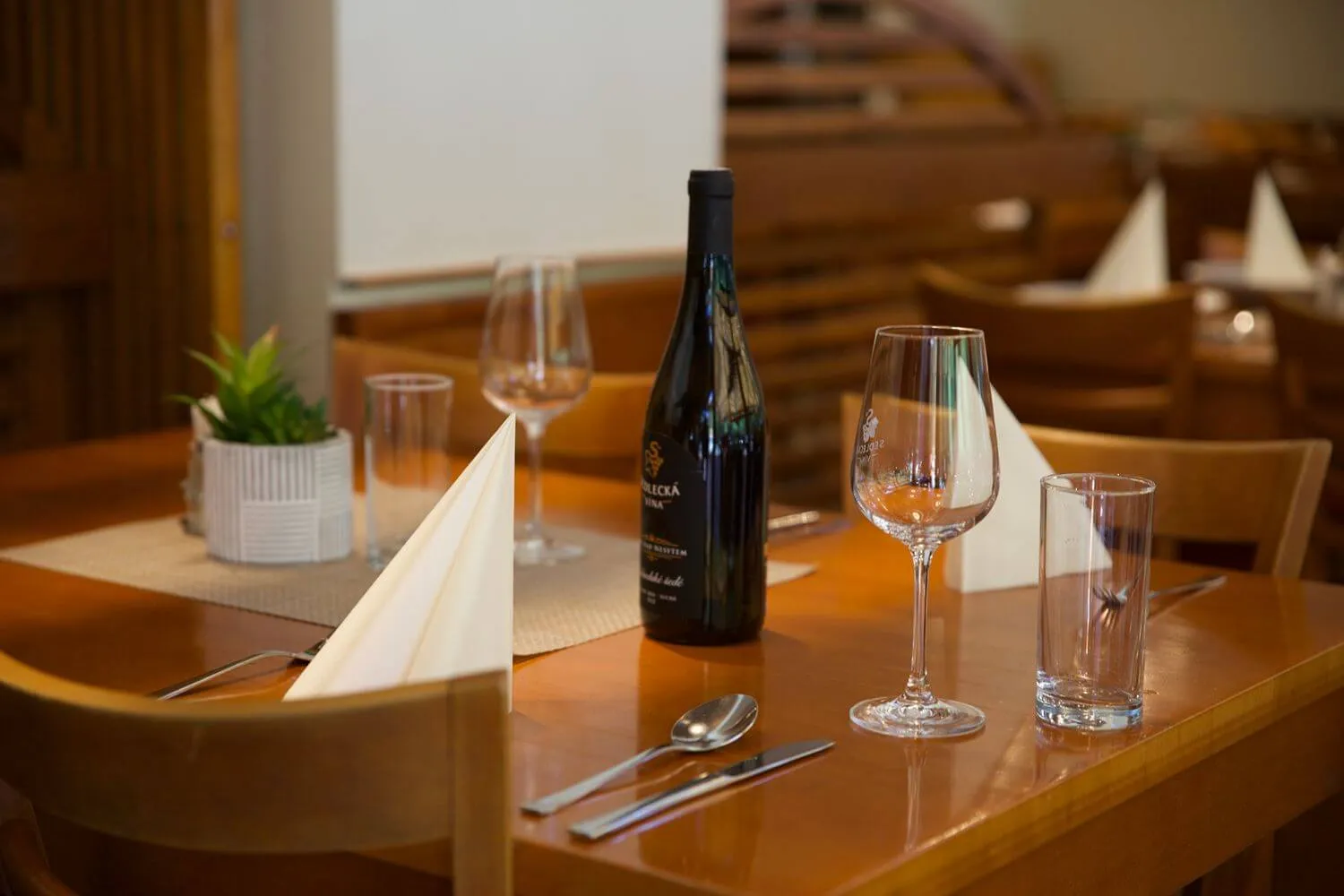 Stůl v restauraci s lahví vína, sklopenými ubrousky, sklenicemi a příbory.