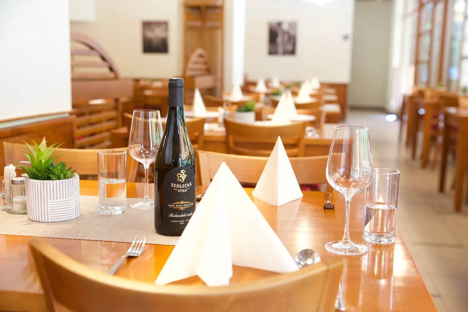 Stůl v restauraci s lahví vína, sklenicemi, bílými ubrousky a malou rostlinou.
