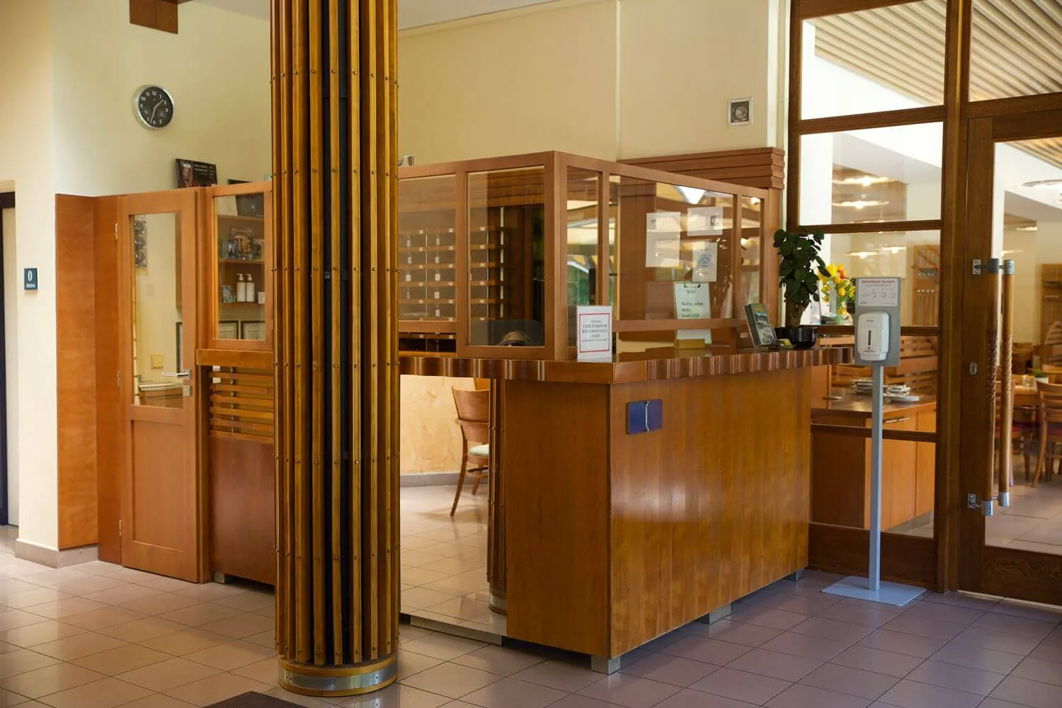 Recepce hotelu ze dřeva s ochrannou přepážkou, stojanem s dezinfekcí a židlí v pozadí.
