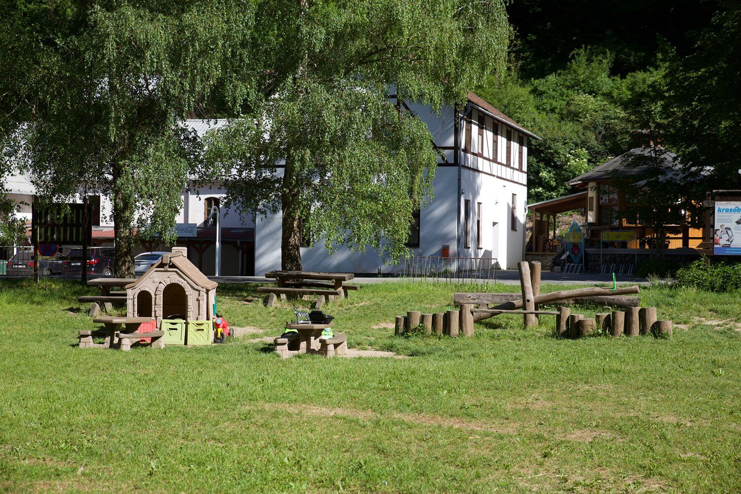 Dětské hřiště s malým domečkem a dřevěným prolézačkovým vybavením na trávníku před domem.