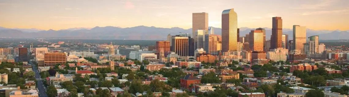 Denver skyline at sunset with downtown skyscrapers, mountains in background, and residential neighborhoods below.