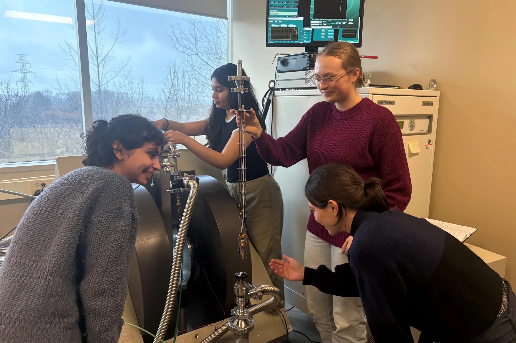 Four researchers examining a sample probe on an EPR spectrometer's electromagnet in a lab, with data on a monitor above.