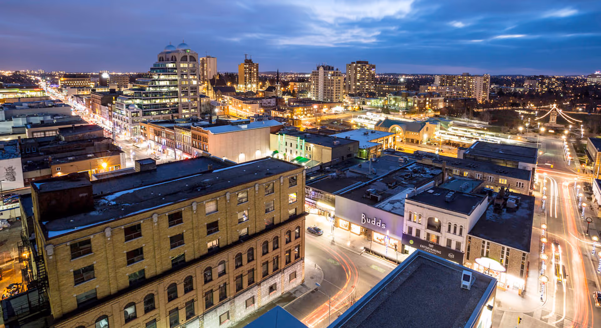 Cityscape at dusk showing buildings, streets with light trails, and a partly cloudy sky.