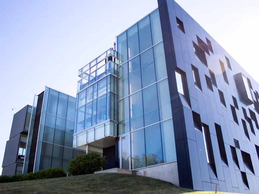 Modern glass and metal building with geometric design on a grassy slope under blue sky.