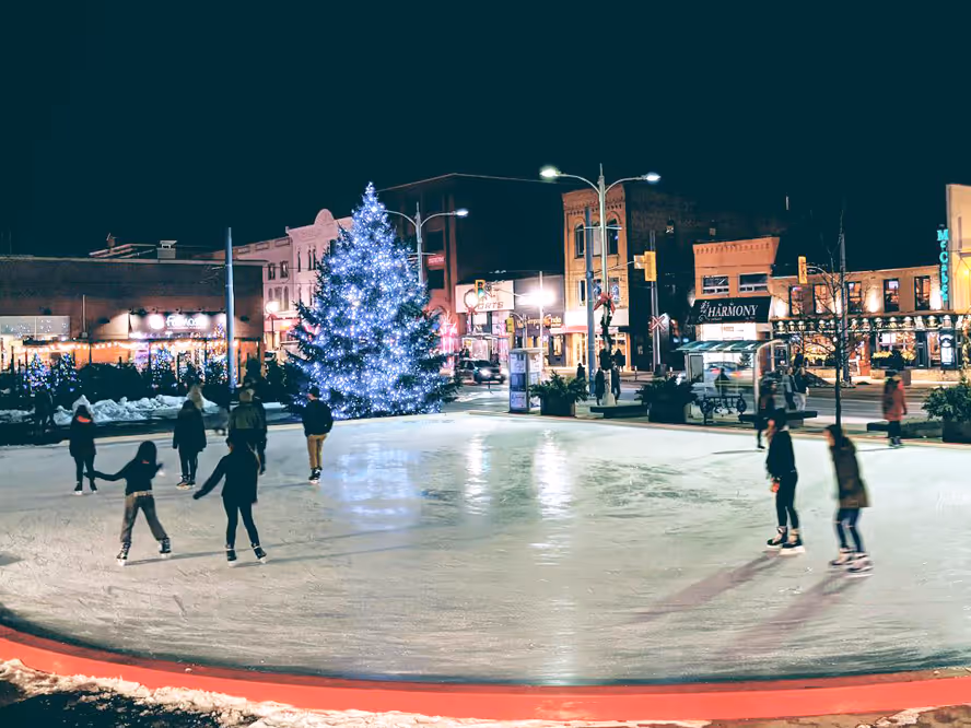 People ice skating at night on a rink near a large Christmas tree with white lights in a town square.