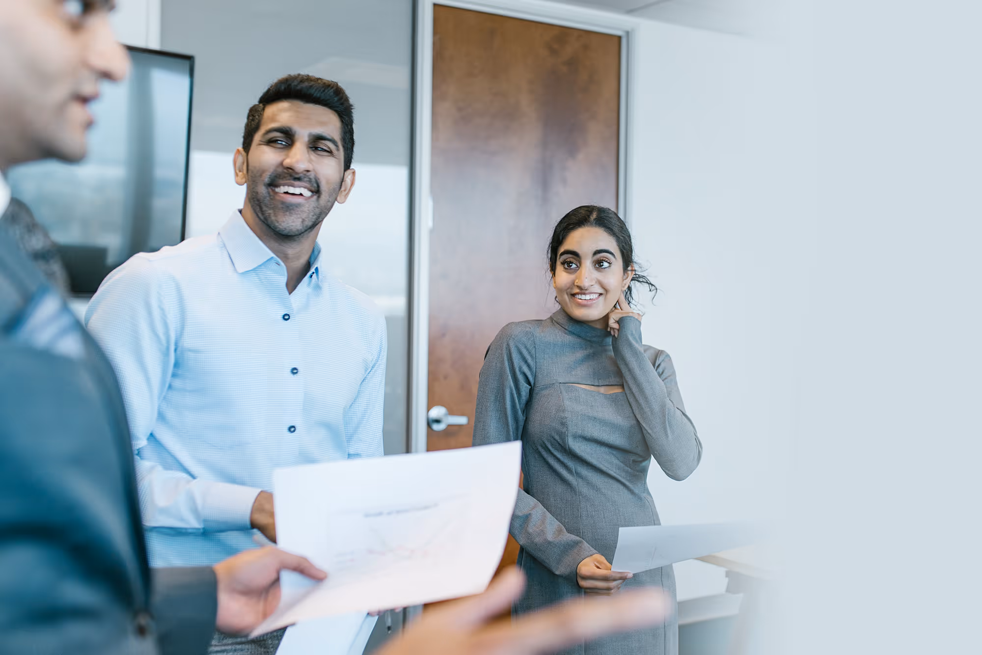 Two smiling professionals listening to a colleague presenting in a modern office meeting room.