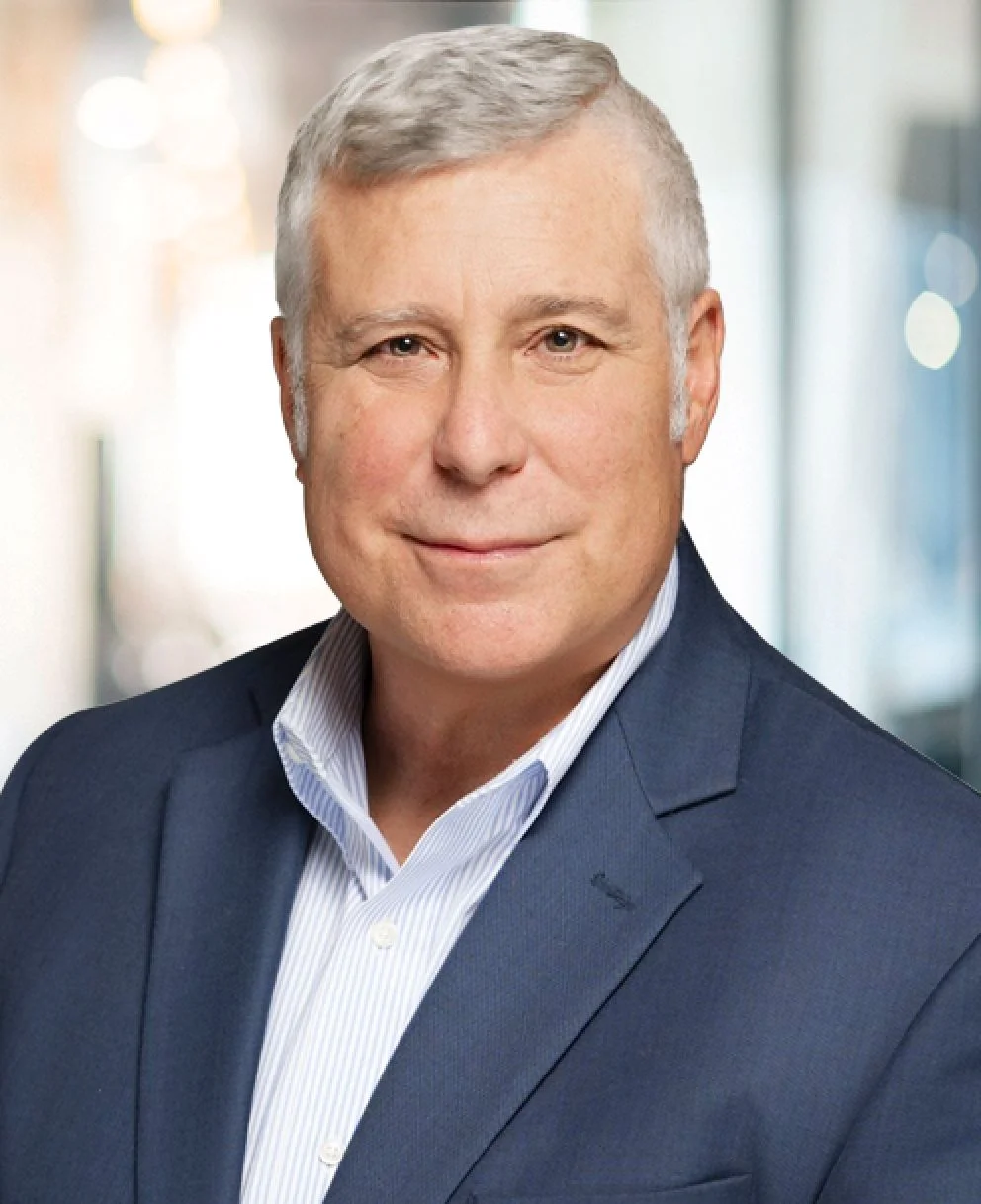 Middle-aged man with gray hair wearing a blue blazer and striped shirt smiling softly.