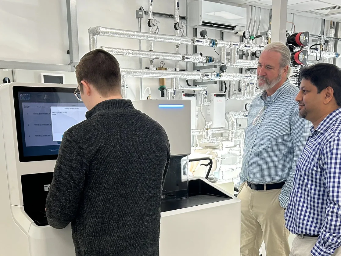 Three men in a lab observing a high-tech spectrometer with exposed piping on the wall.