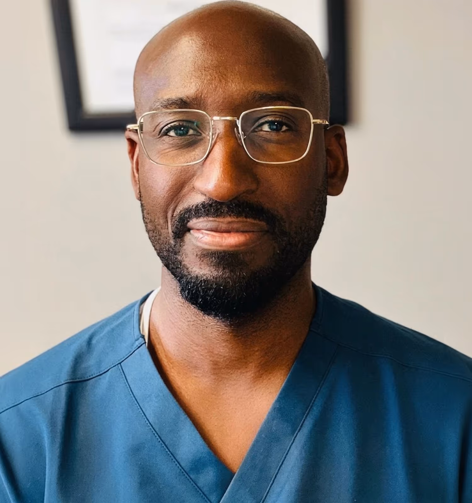 Portrait of a smiling bald man with glasses and a beard wearing blue medical scrubs.
