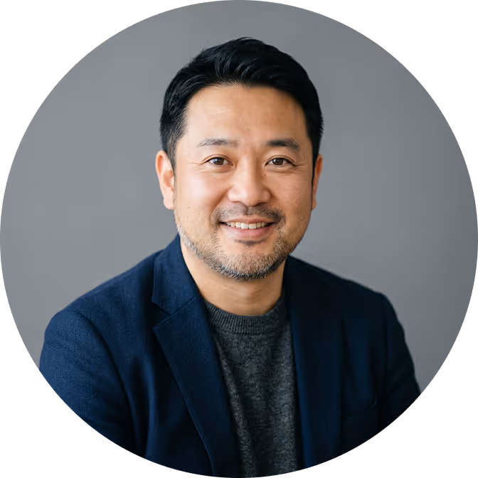 Smiling middle-aged Asian man with short black hair and beard wearing a dark blazer and gray sweater in front of a gray background.