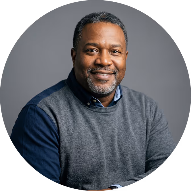 Smiling middle-aged man with short curly hair and salt-and-pepper beard wearing a grey sweater over a blue collared shirt against a grey background.
