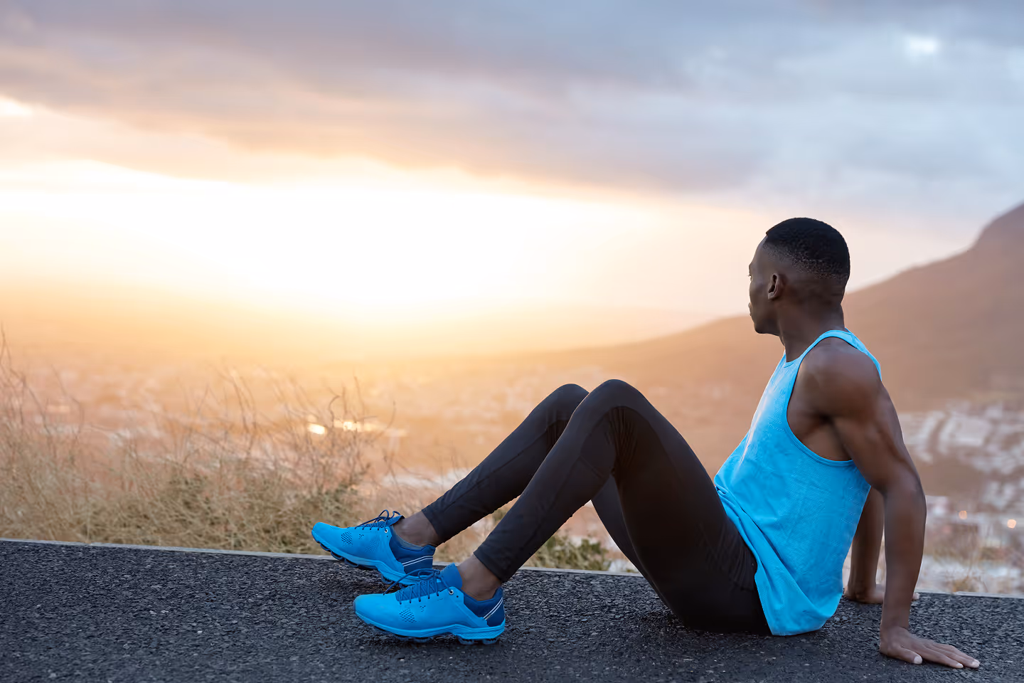 Athletic man in blue tank top and running shoes sitting on a roadside, watching a bright sunset over distant hills.