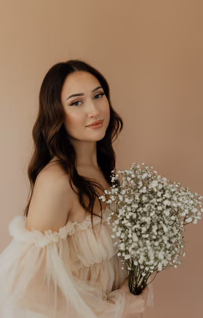 Young woman with long dark hair in light off-shoulder dress holding a bouquet of small white flowers.