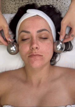 Woman with eyes closed receiving facial massage with metal roller balls on cheeks.