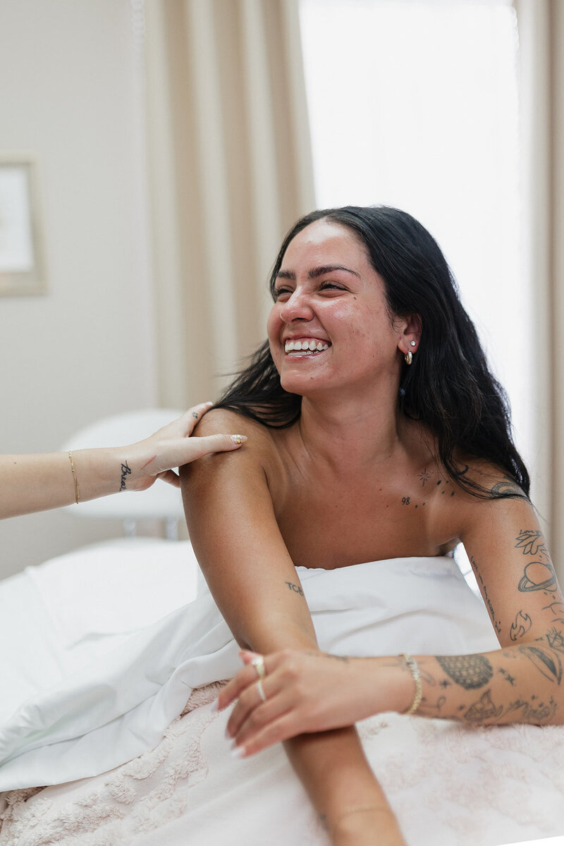 Smiling tattooed woman with long dark hair lying on a bed covered with a white blanket.