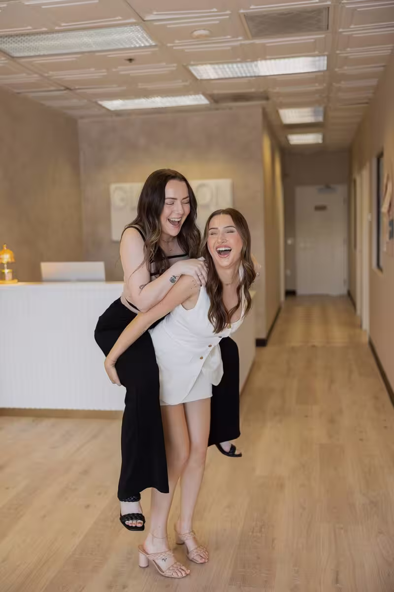 Two women laughing, one giving the other a piggyback ride in a modern indoor hallway with wooden floor.