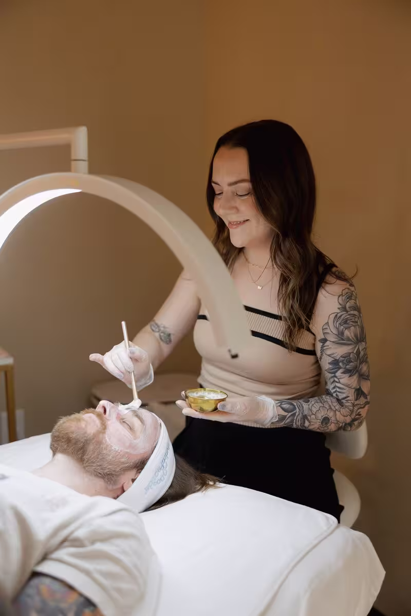 Tattooed esthetician applying a facial treatment to a man's face under a ring light.